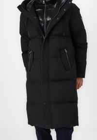 Lång svart vadderad vinterjacka med dragkedja och tryckknappar, med fickor med dragkedja och klaff, buren över en glänsande svart innerjacka.