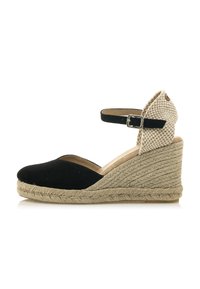Sandalía de cuña de ante negro con una correa de tobillo tejida en beige, que presenta un tacón de cuerda de yute natural y un diseño de punta cerrada.