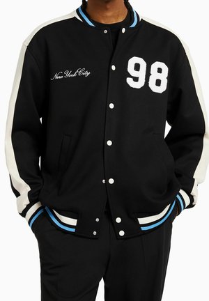 Zwarte varsityjas met witte strepen op de mouwen, "98" patch op de borst, "New York City" geborduurd, blauw-witte gestreepte manchetten, gedragen met handen in de zakken.