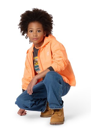 Veste à capuche légère orange vif, t-shirt graphique gris en dessous, jean en denim bleu et bottes marron clair avec des lacets jaunes.