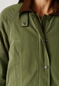 Sieviete, kas ģērbjas olīvu zaļā jaka ar brūnu korduroja apkakli, metāla spiedpogu pogām un redzamu sudraba rāvējslēdzēju.