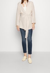 Beige omslagströja med långa ärmar och knytbälte, tillsammans med mörka skinnyjeans och ljusgula klackskor, stående mot en neutral bakgrund.