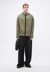 Olivovo zelená mikina na zips s čiernym zipsom, nosená cez voľné čierne nohavice, pravou rukou drží čiernu tašku. Minimalistický dizajn.