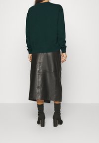 Persona che indossa un top a maniche lunghe verde scuro, una gonna midi in pelle nera e stivali alla caviglia con tacco neri, in piedi contro uno sfondo chiaro e semplice.