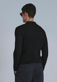 Haut noir à manches longues côtelé avec un col, porté avec un pantalon gris. Modèle portant des lunettes de soleil sombres, vue de dos.