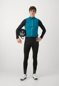 Radsportoutfit mit einer petrolfarbenen Weste mit Reißverschluss, schwarzer langärmeliger Basisschicht und schwarzen Strumpfhosen. Ein schwarzer Helm wird in einer Hand gehalten.