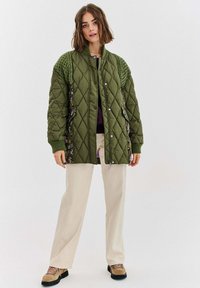 Žena v zelené prošívané bundě s pletenými rameny, béžových kalhotách s širokými nohavicemi a hnědých turistických botách, stojící na jednobarevném bílém pozadí.