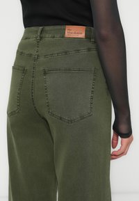 Olivově zelené džíny z denimu mají koženou etiketu na pase. Zadní kapsy mají jednoduchý design s viditelným švem.