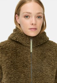 Bruine fleece jas met een hoge kraag, voorzien van een ritssluiting aan de voorkant en een textuur van fluffy stof. Subtiele glans op de ritshardware.