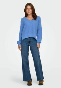 Blauwe gebreide trui met V-hals en een gestructureerd patroon, gecombineerd met hoge taille jeans met wijde pijpen. Bruine enkellaarzen maken de outfit compleet.