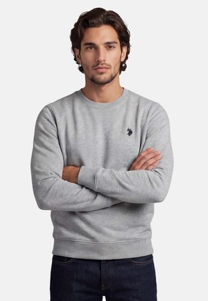 Man with dark hair and beard wearing a light grey sweatshirt with a small dark logo, standing with arms crossed against a plain background.