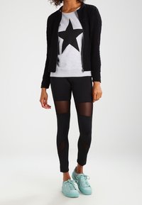 Chaqueta negra con cremallera sobre un sudadera gris con una estrella negra. Leggings negros con secciones de malla. Zapatillas deportivas verde menta.