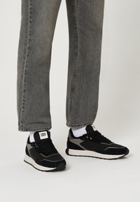 Sneaker neri e grigi con accenti in camoscio e feltro, suola bianca e superfici testurizzate, indossati con jeans grigi e calzini bianchi.