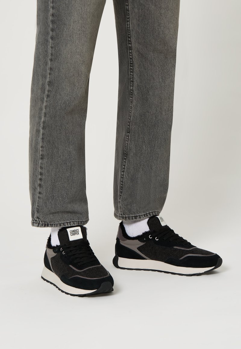 Sneaker neri e grigi con accenti in camoscio e feltro, suola bianca e superfici testurizzate, indossati con jeans grigi e calzini bianchi.