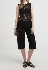 Top senza maniche in pizzo nero sovrapposto a un tessuto nero solido. Abbinato a pantaloni cropped neri e sandali neri senza chiusura. Clutch minimalista in mano.