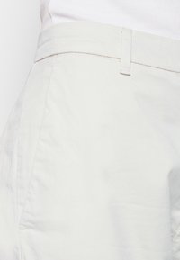 Pantalon de couleur claire en mélange de coton, présentant une texture lisse, des poches latérales et une ceinture plate avec une boucle de fixation discrète.