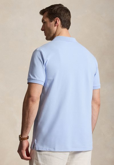 Άνδρας που φοράει ανοιχτό μπλε μπλούζα πόλο με κοντά μανίκια και μπεζ παντελόνι, βλέπεται από πίσω σε απλό φόντο.