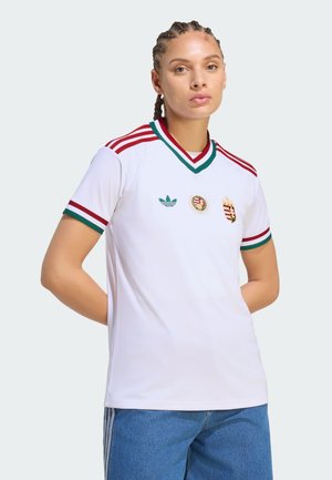 Jongere met gevlochten haar draagt een wit sportshirt met rode en groene strepen en emblemen, gecombineerd met blauwe spijkerbroek, tegen een effen achtergrond.