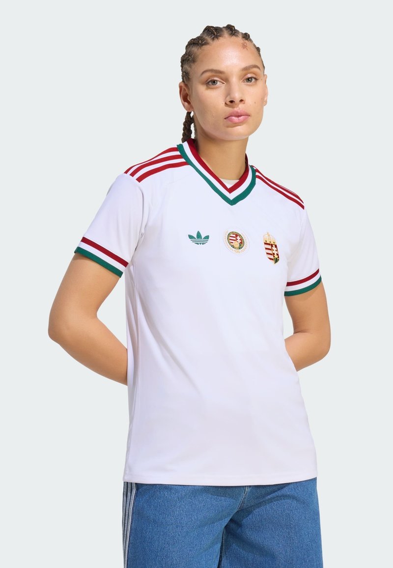 Jeune personne aux cheveux tressés portant un maillot de sport blanc avec des rayures rouges et vertes ainsi que des emblèmes, associée à un pantalon en denim bleu, sur un fond uni.