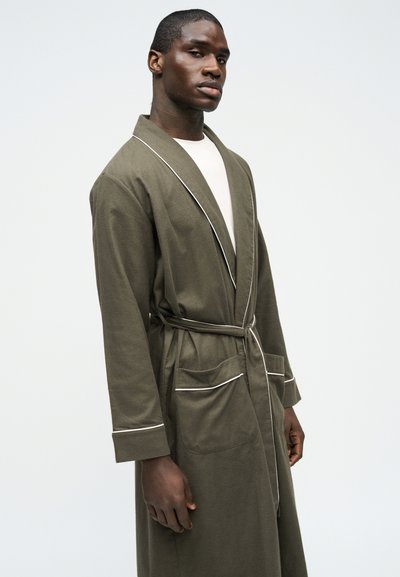 Άνδρας που φορά μακριά ρόμπα ελιάς με λευκή ρίγα, δεμένη στη μέση, πάνω από λευκό πουκάμισο, στέκεται μπροστά σε απλό ανοιχτόχρωμο φόντο.