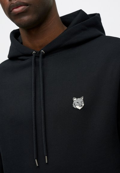 Haut à capuche noir en tissu texturé avec une broderie de tête de renard argenté sur la poitrine. Comprend un cordon de serrage ajustable avec embouts en métal.