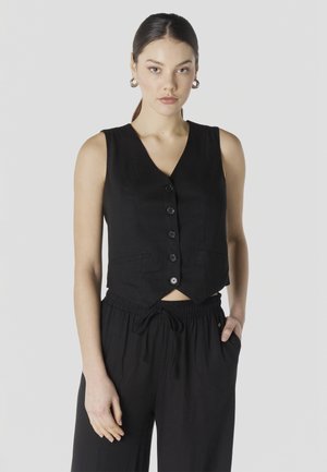 Junge Frau mit dunklem Haar im Pferdeschwanz trägt ärmellose schwarze, geknöpfte Weste und schwarze Hose mit Kordelzug, steht mit einer Hand in der Tasche.