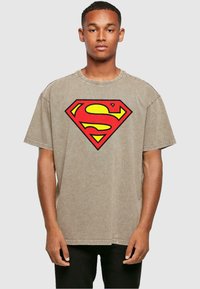 Camiseta de algodón beige con un gran logo de Superman en rojo y amarillo en el pecho, con mangas cortas y escote redondo.