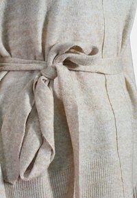 Cardigan beige lavorato a maglia con una cintura in tessuto legata in vita. La trama è morbida, con un leggero motivo melange e polsini a coste.