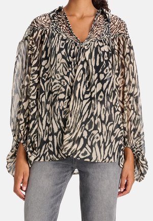 Kvinde iført gennemsigtig bluse med lange ærmer med sort og beige abstrakt dyreprint, kombineret med lysegrå jeans, stående mod en hvid baggrund.