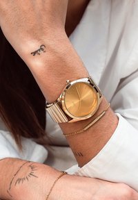 Montre en or avec un cadran rond, un bracelet métallique et un design minimaliste. Elle présente un cadran doré et une texture lisse. Accent de bracelet avec de petites pierres.