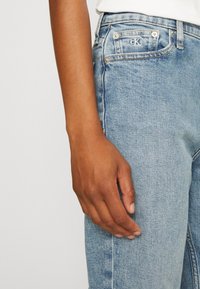 Vaaleansiniset farkut, joissa on näkyvät saumat, etutasku, jossa on pieni logo, ja suora lahje. Pehmeä rakenne ja klassinen muotoilu.