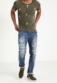 Oliivivihreä T-paita, jossa on valkoisia ristikkokuvioita, kuluneet siniset farkut ja vihreät lenkkarit. Rentoa istuvuutta, jossa on käännetyt hihansuut ja kontrastiväriset tikkaus.