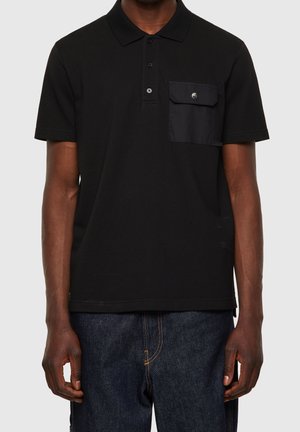 Homme portant un polo noir à manches courtes avec trois boutons et une poche à rabat sur la poitrine gauche, associé à un jean bleu foncé.