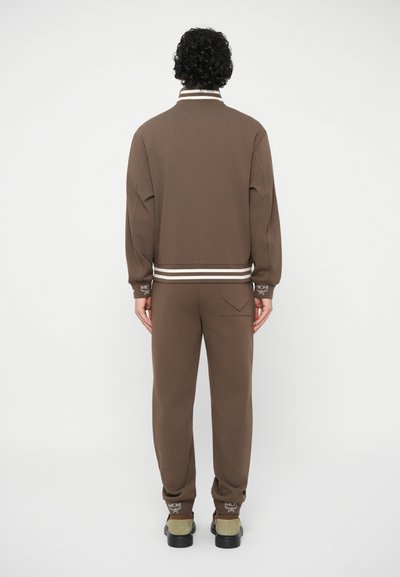 Pessoa de pé, de costas, vestindo um fato de treino castanho com punhos e colarinho riscas, logótipos bordados e sapatos bege e pretos.