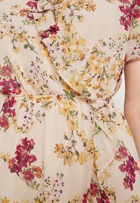 Puvun kukkakuosi beigen värisessä kankaassa, jossa on vaaleanpunaisia, keltaisia ja vihreitä kuvioita. Röyhelödetaljit kaula-aukossa ja vyötäröllä elastisella keräämisellä.