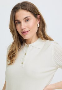 Femme aux cheveux châtain clair porte un polo blanc en maille à manches courtes et des boucles d'oreilles argentées en forme d'anneaux, posant devant un fond clair uni.
