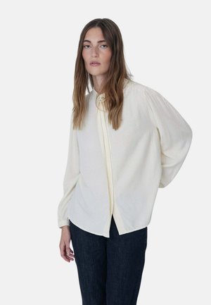 Mujer con cabello largo y castaño viste una blusa blanca suelta con botones y pantalones oscuros, de pie frente a un fondo claro y liso.