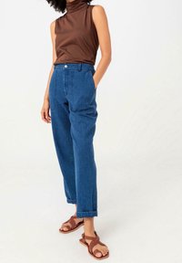 Brown sleeveless top paired with blue loose-fit denim trousers. Sandals feature crisscross straps. Smooth fabric with a clean design.