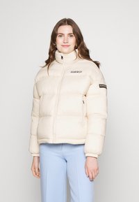 Crème pufferjack met een hoge kraag, horizontale gestikte secties en zwarte logoaccenten op de borst en de mouw. Zachte textuur.