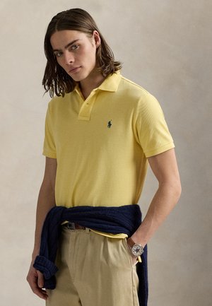 Jeune homme aux cheveux mi-longs portant un polo jaune, un pantalon beige, un pull bleu marine noué autour de la taille et une montre-bracelet, debout devant un fond uni.