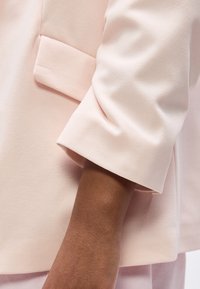 Χέρι με μανίκι ελαφρώς ροζ σακακιού με λεπτομέρεια τσέπης με καπάκι κοντά στον καρπό, σε φωτεινό φόντο.