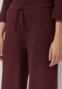 Bordeaux gekleurde stoffen broek met een gekreukeld materiaal, voorzien van een elastische tailleband en een koord voor verstelbaarheid.