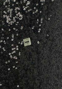 Tissu noir à sequins avec de petits sequins ronds et une étiquette argentée portant la mention "Manila Grace", mettant en valeur une surface brillante et texturée.