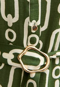 Groene stof met wit abstract kettingpatroon, met een gouden gesp in onregelmatige vorm en een kleine ronde knoop.