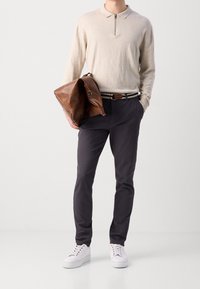 Hellbeiges Langarm-Poloshirt mit Reißverschluss, kombiniert mit dunkelgrauer Hose und weißen Turnschuhen. Eine braune Lederhandtasche wird in der linken Hand gehalten.