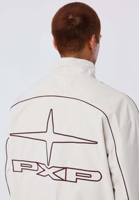 Persoon met kort bruin haar, draagt een wit jasje met zwarte bies en een groot PXG-logo op de achterkant.