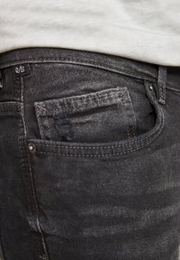 Vaqueros de mezclilla gris oscuro con una superficie texturizada, que cuentan con un bolsillo trasero y una pequeña etiqueta con "Q/S" cosida cerca de la cintura.