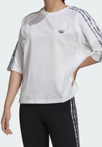 Vit kortärmad sweatshirt med blommönstrad sidrand, rund halsringning och sidodragband för justerbar passform. Adidas-logotyp på bröstet.