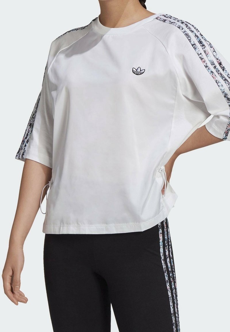 Vit kortärmad sweatshirt med blommönstrad sidrand, rund halsringning och sidodragband för justerbar passform. Adidas-logotyp på bröstet.
