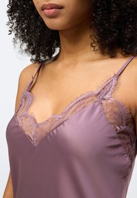 Γυναίκα με αμάνικο τοπ από σατέν σε χρώμα μαβί, με λεπτή δαντέλα κατά μήκος του ντεκολτέ και λεπτές τιράντες στους ώμους.